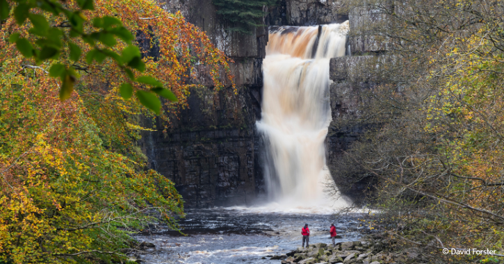 Two people stood at the foot of High Force Waterfall during autumn.